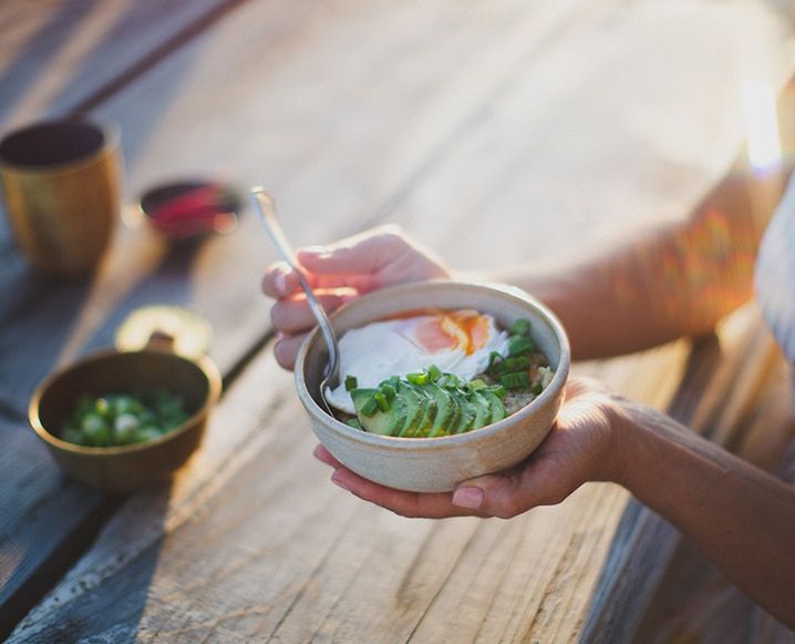 Hands holding a warm bowl of Conjo Congee in morning light topped with egg, avocado and greens - Photo by The Chalk Board Mag / Maui Maka